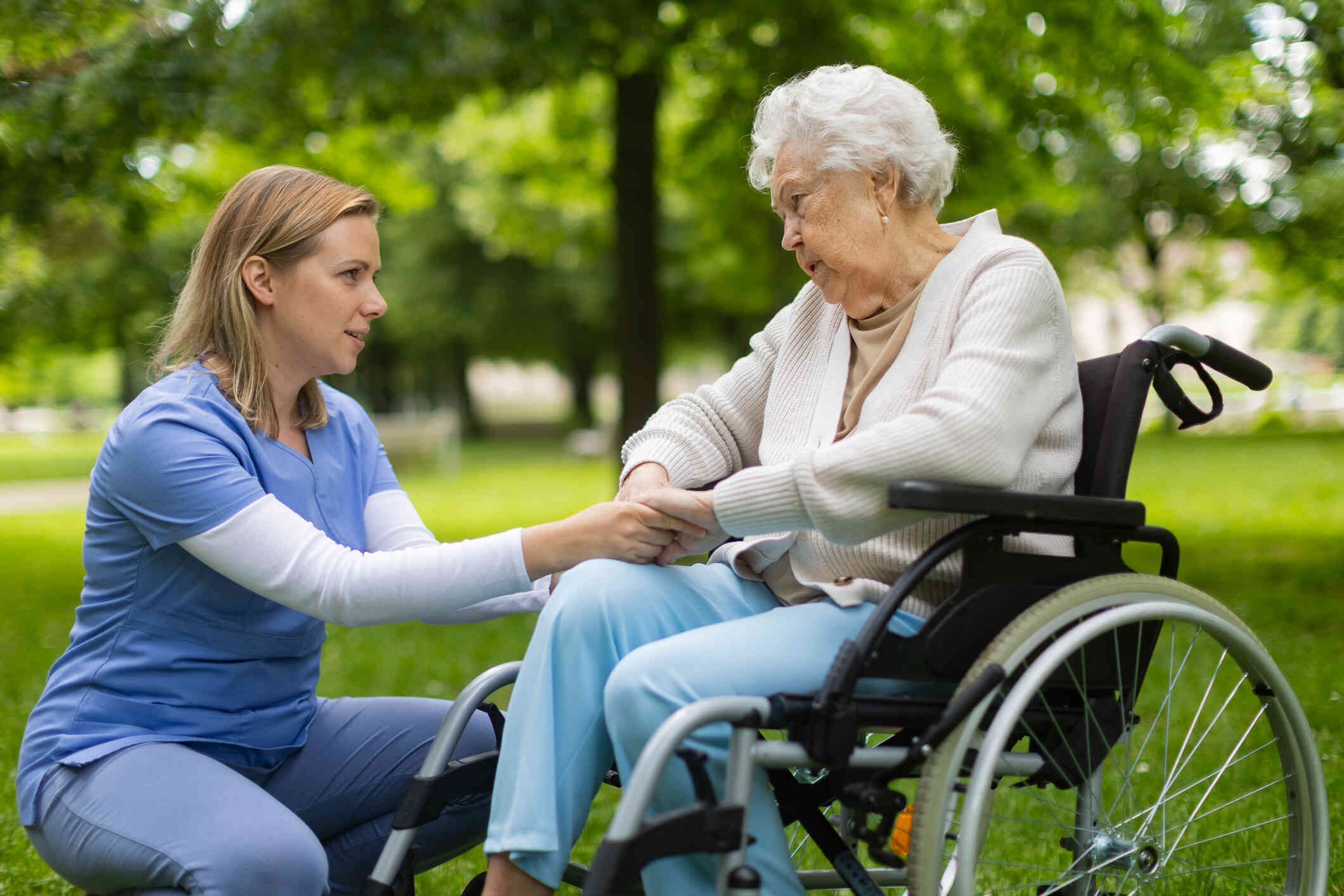 An elderly woman sits in a wheelchair outside on a sunny day as her female nurse squats down infront of her and holds her hands.